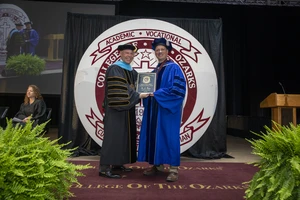 This image depicts two men in graduation robes and regalia. They are standing on a stage with greenery to each side and a large logo of College of the Ozarks as the backdrop. The men are smiling at the camera with an award between them. 